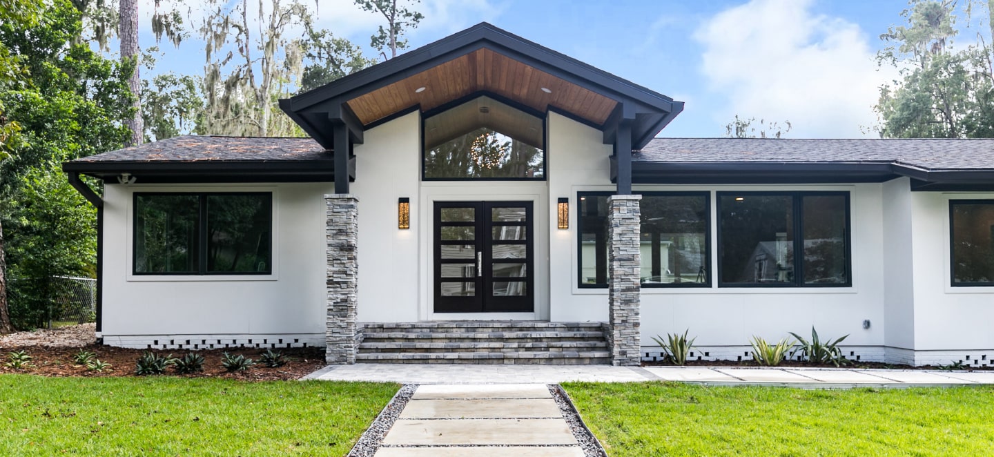 Modern home exterior with a welcoming entryway featuring stone columns, a large glass front door, and wide steps leading to a pathway, surrounded by a green lawn and landscaping.
