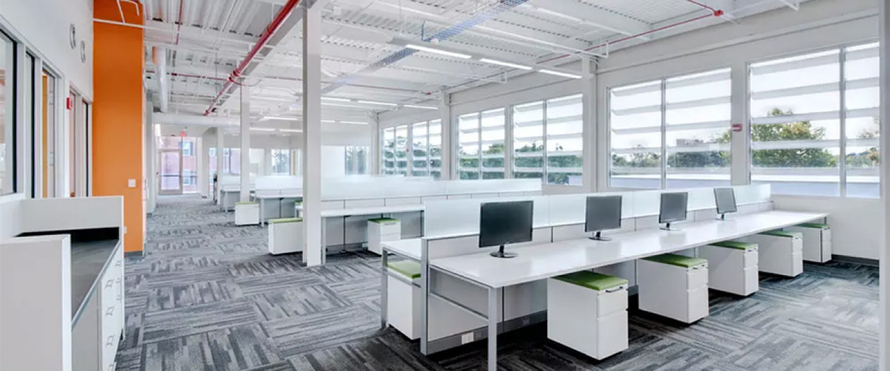 A modern, brightly lit open-plan office with rows of white desks, computer monitors, green-accented chairs, and large windows letting in natural light. The space features gray patterned carpet and exposed ceilings.