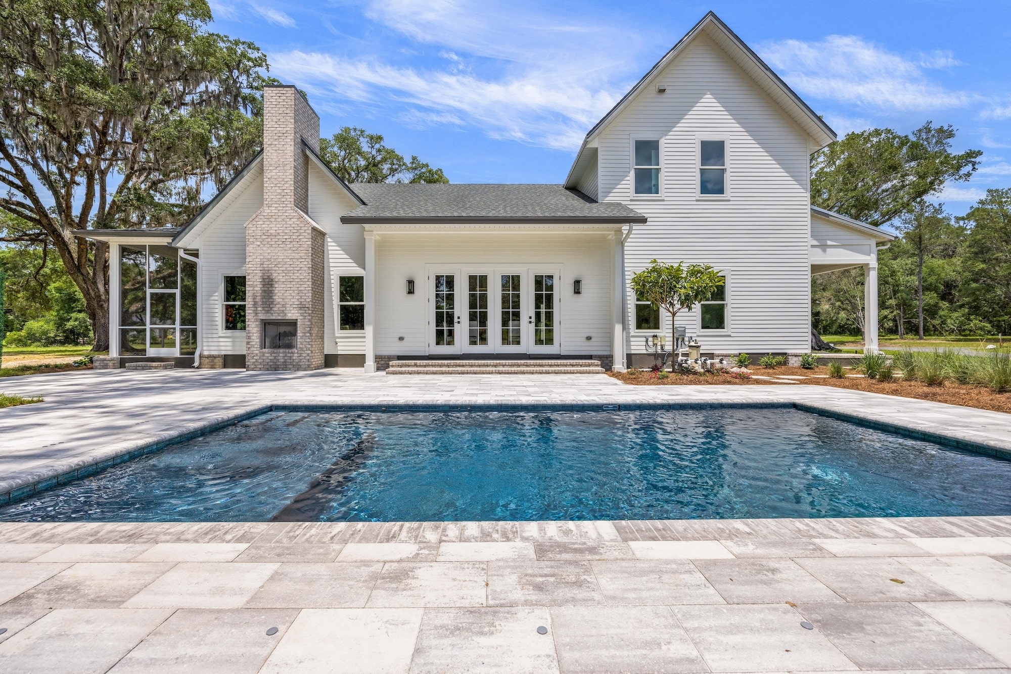 A white farm house sits behind a white stone patio and in-ground pool