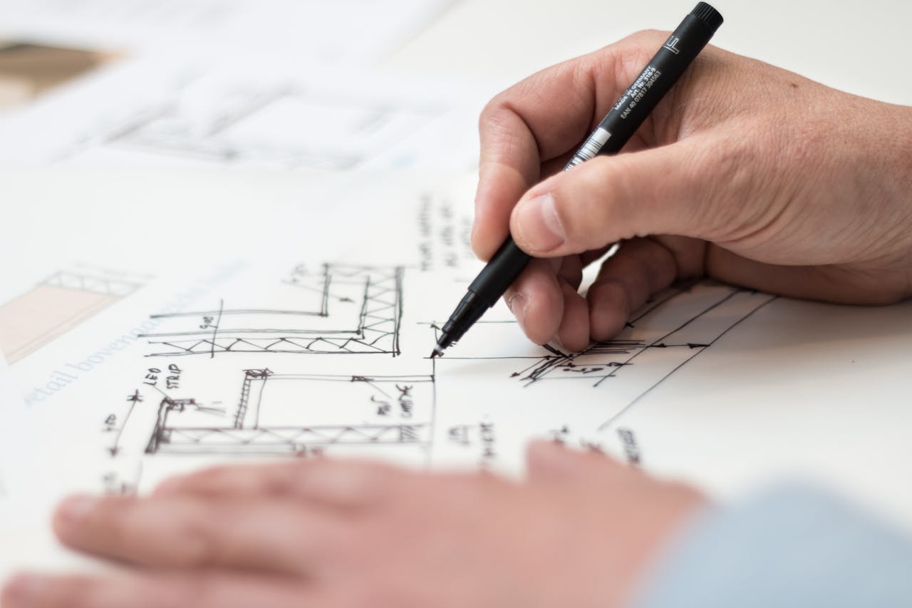 A person is drawing architectural plans with a black pen on white paper, showing detailed building sketches and measurements. Another hand steadies the paper while the person works.